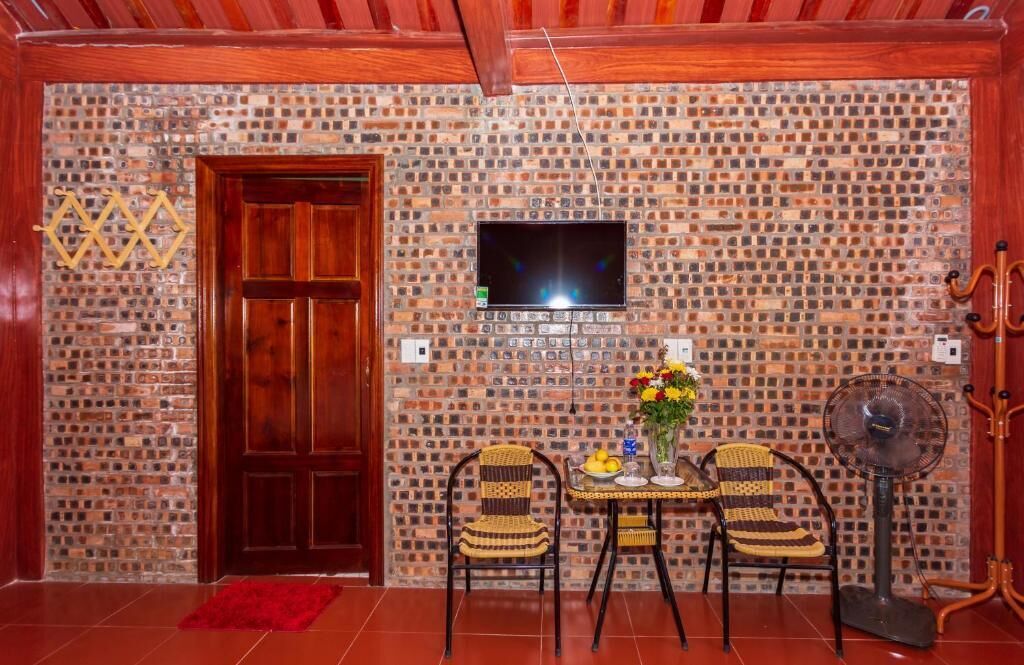 Quarto com parede de tijolos, porta de madeira, televisão na parede, duas cadeiras de vime, mesa pequena com um vaso de flores e um ventilador.
