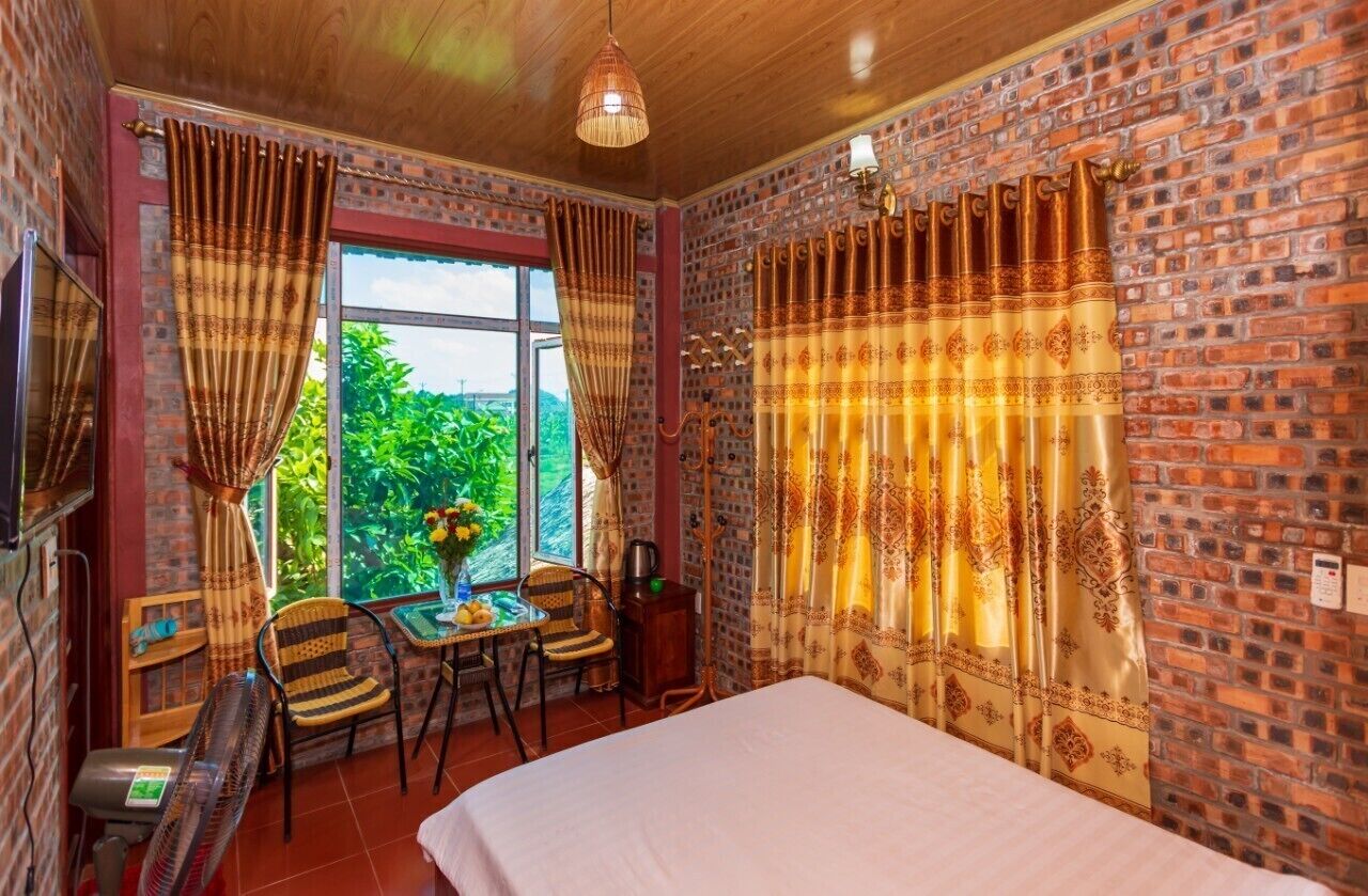 Una habitación con cama, dos sillas, una mesita con un jarrón y una ventana con cortinas.