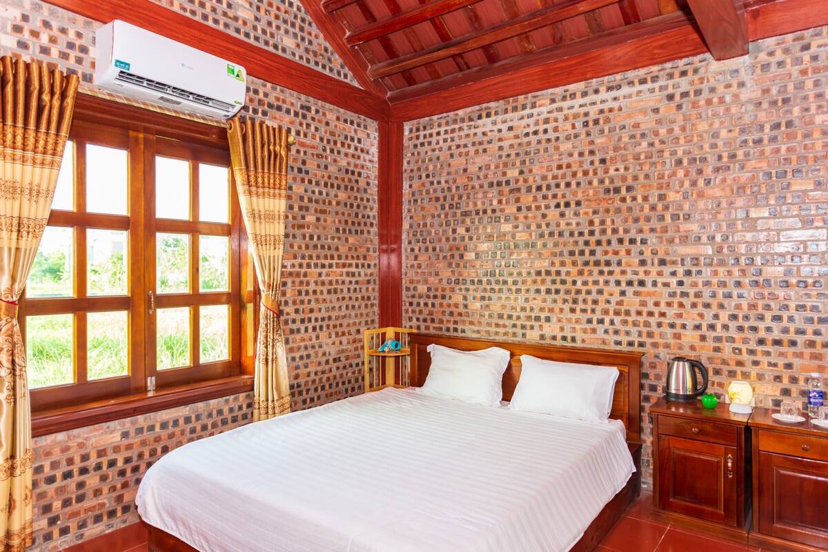 Une pièce avec un mur dont le motif est en briques, un lit en bois avec une literie blanche, une fenêtre avec des rideaux et une petite table de chevet en bois sur laquelle se trouve une bouilloire.