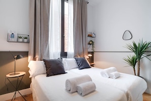 Un lit bien fait, avec du linge de lit blanc et des serviettes pliées, une lampe de chevet, une petite table ronde et une plante en pot.