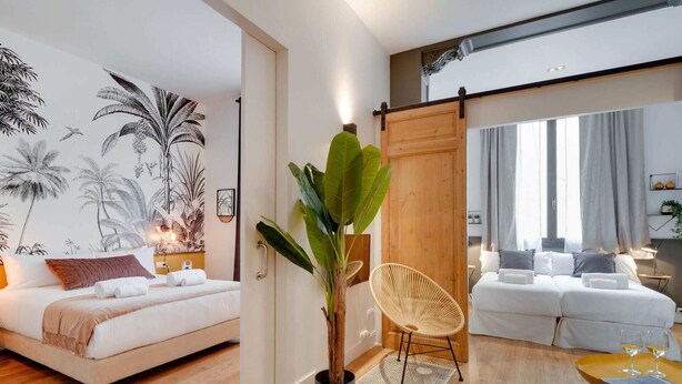 A modern hotel room with a bed, a sitting area with a chair, a large potted plant, and a view of the city through large windows. - Sweett | Sepulveda 3B (Barcelona)