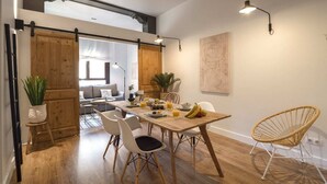 Een eetruimte met een houten tafel, witte stoelen en een rieten stoel. Achtergrond heeft een bank en een plant.