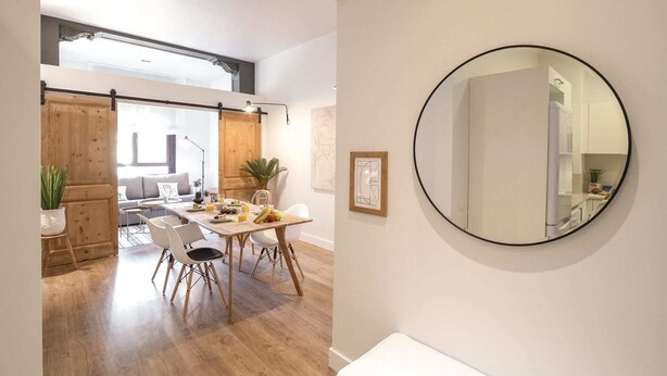 Een moderne eethoek met een houten tafel en stoelen, een grote ronde spiegel aan de muur, en uitzicht op een woonruimte met een bank en een plant.