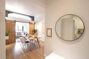 A modern dining area with a wooden table and chairs, a large round mirror on the wall, and a view into a living space with a sofa and a plant.