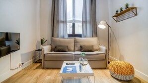 Een moderne woonkamer met een beige bank, een salontafel met afstandsbedieningen en glazen, een vloerlamp en een klein plantenplankje.