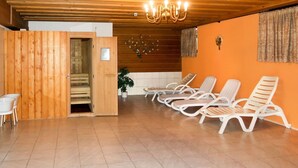 A sauna room with wooden walls, a wooden ceiling, and a chandelier. There are white lounge chairs and a small table with a chair.