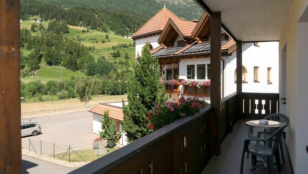 Un balcón con vistas a un paisaje montañoso, un edificio con techo inclinado y una mesa con sillas.