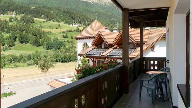 Un balcón con vistas a un paisaje montañoso, un comedor con sillas y un edificio de techo con tejas.