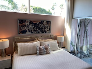 Ein Schlafzimmer mit einem großen Bett, zwei Nachttischlampen, einem Bild an der Wand und Blick auf Bäume durch ein Fenster.