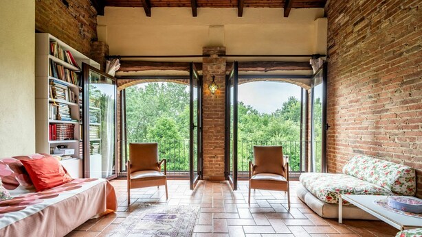 Une pièce chaleureuse avec un mur en briques, une bibliothèque, un canapé, deux fauteuils et une grande fenêtre donnant sur les arbres.