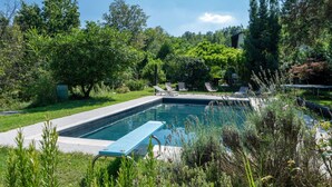 Une piscine rectangulaire aux bords bleus, entourée d’une végétation luxuriante et d’arbres. On y trouve des chaises longues et une petite table près de la piscine.