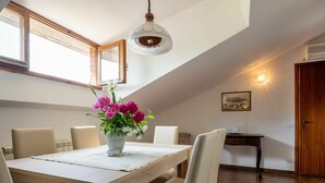 A dining area with a wooden table, six chairs, a vase of flowers, a pendant light, a painting on the wall, and a door with an air conditioning unit. - Nice apartment for 4 people with WIFI, A/C, pool, TV and patio (Castagnole Delle Lanze)