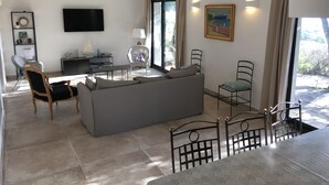 Ein modernes Wohnzimmer mit grauem Sofa, einem Esstisch und Stühlen. Ein Flachbildfernseher ist an der Wand montiert, ein kleiner Beistelltisch mit Lampe steht daneben, und über dem Fernseher hängt ein Gemälde.