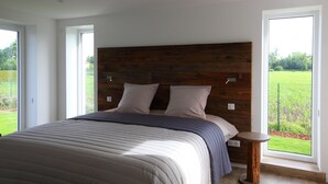 A modern bedroom with a large bed, wooden headboard, and a view of a green field through a large window.