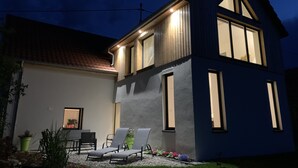 A two-story house with a sloped roof, a balcony, and a well-lit front porch with outdoor seating.