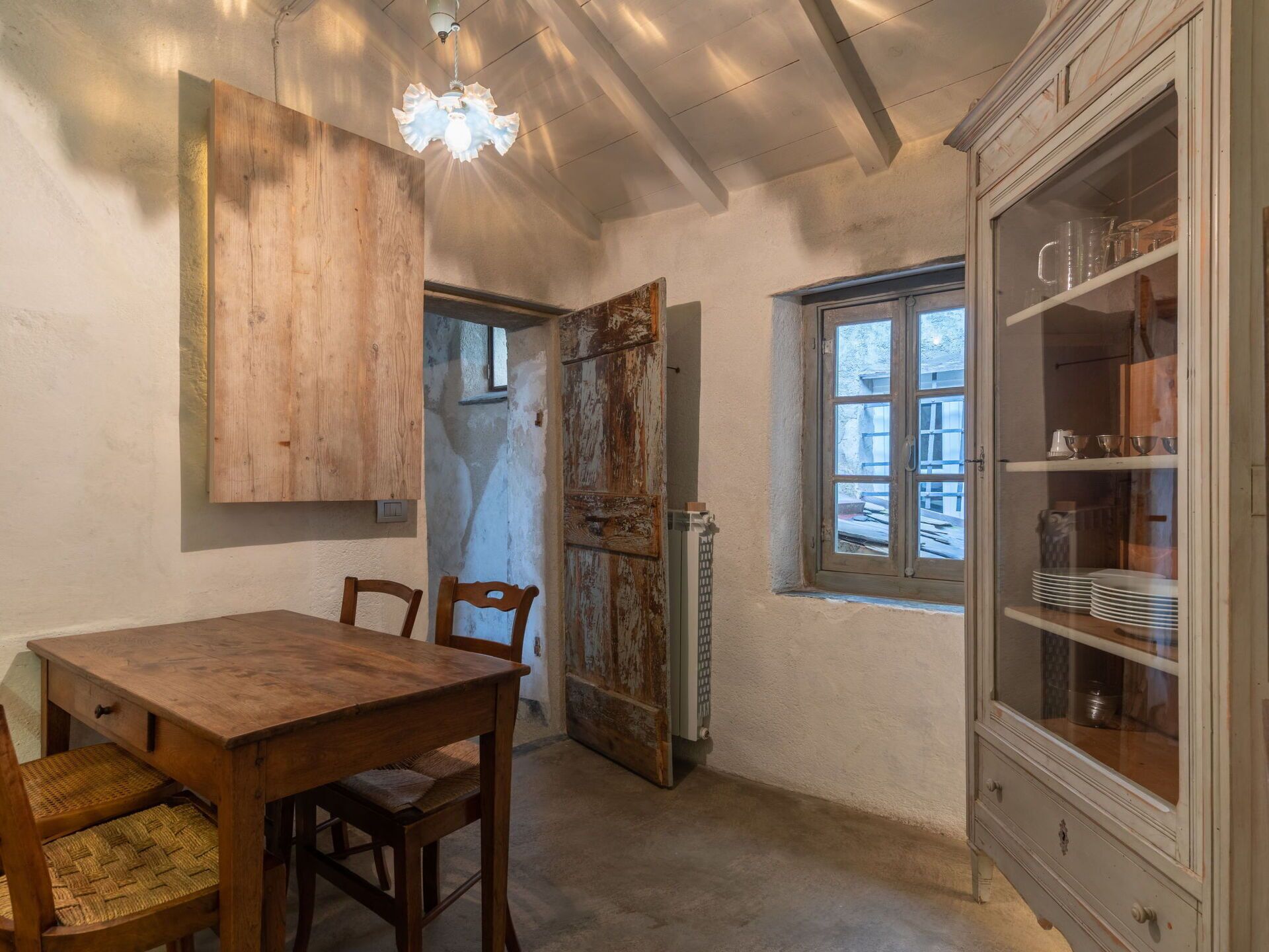 A rustic dining area with a wooden table and chairs, a cabinet with glass doors, and a window with a view of the outside.