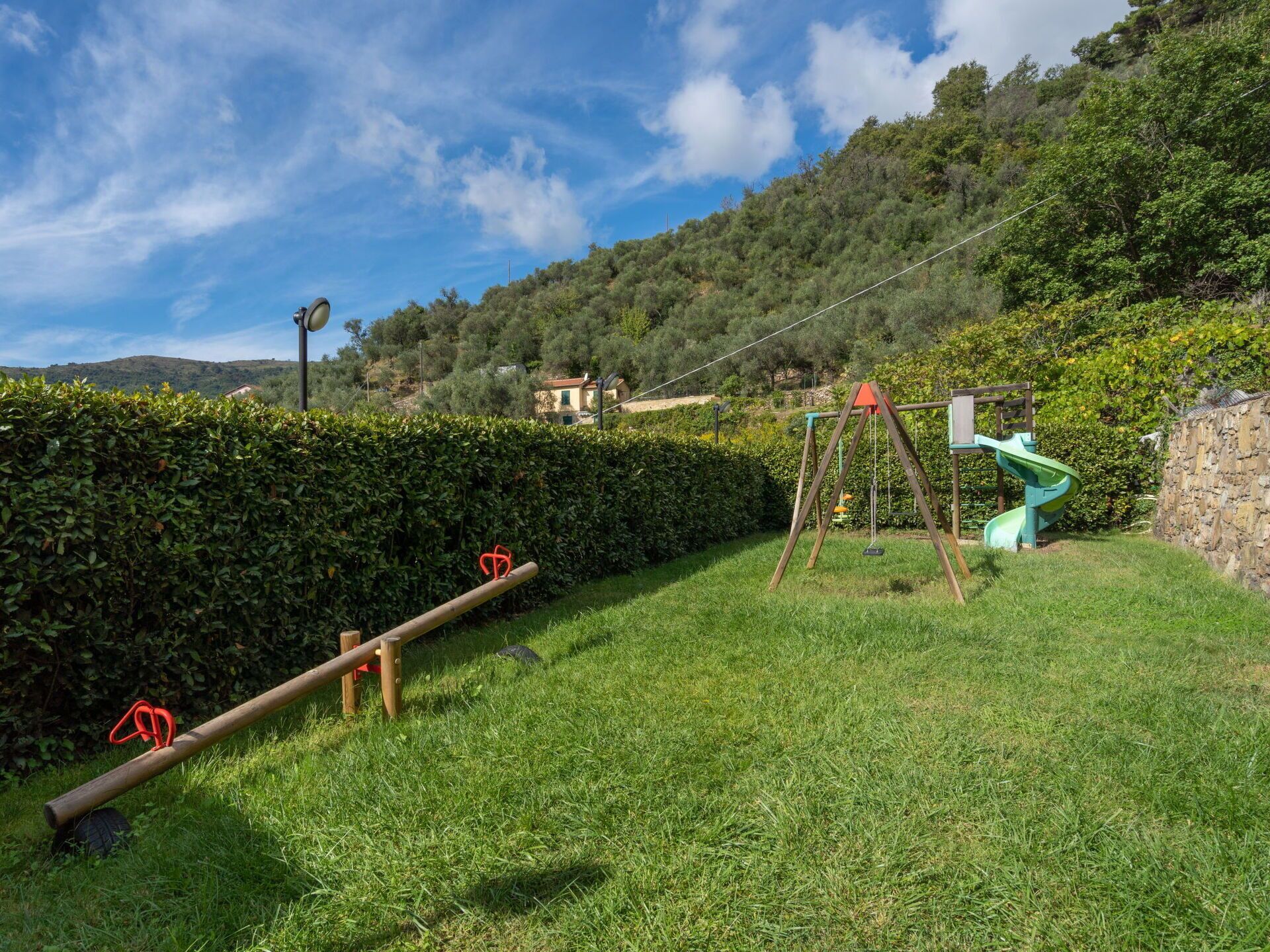 A grassy outdoor area with a swing set and a slide, surrounded by a hedge and a stone wall.
