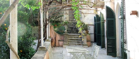 A covered outdoor seating area with metal tables and chairs, surrounded by greenery and a stone wall.