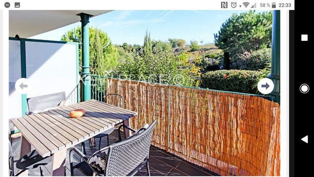 Una mesa y sillas de madera en un balcón con cerca de bambú, con vista a un jardín con diversas plantas y árboles.