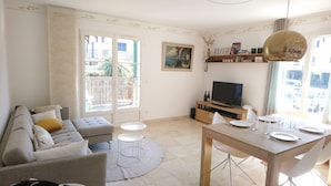 Una sala de estar con un sofá, una mesa de comedor con sillas, un televisor y un balcón con vistas a edificios y zonas verdes.