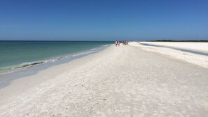 Une vaste plage de sable fin, bordée d’une eau d’un bleu limpide, où quelques personnes se promènent le long du rivage.