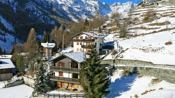 Ein mehrstöckiges Chalet mit Balkon, umgeben von schneebedeckten Bergen und Bäumen.