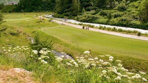 Un parcours de golf avec une rivière, des montagnes et un chalet en arrière-plan.