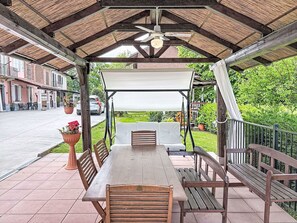 En overdækket terrasse med et bord og bænke, en ventilator i loftet samt udsigt til en gade med bygninger og grønt.