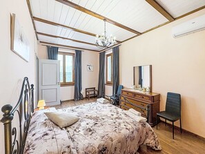 Et soveværelse med seng, kommode, stol, skrivebord, spejl, lysekrone og et vindue med gardiner.