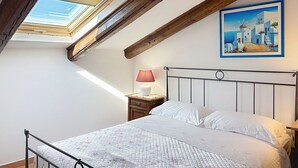 Een slaapkamer met dakraam, een bed met witte beddengoed, een nachtkastje met een lamp en een schilderij van een kustlandschap.