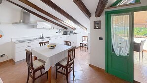 Een keuken met witte kastjes, houten balken, een eettafel met stoelen en een groene deur die leidt naar een buitenruimte.