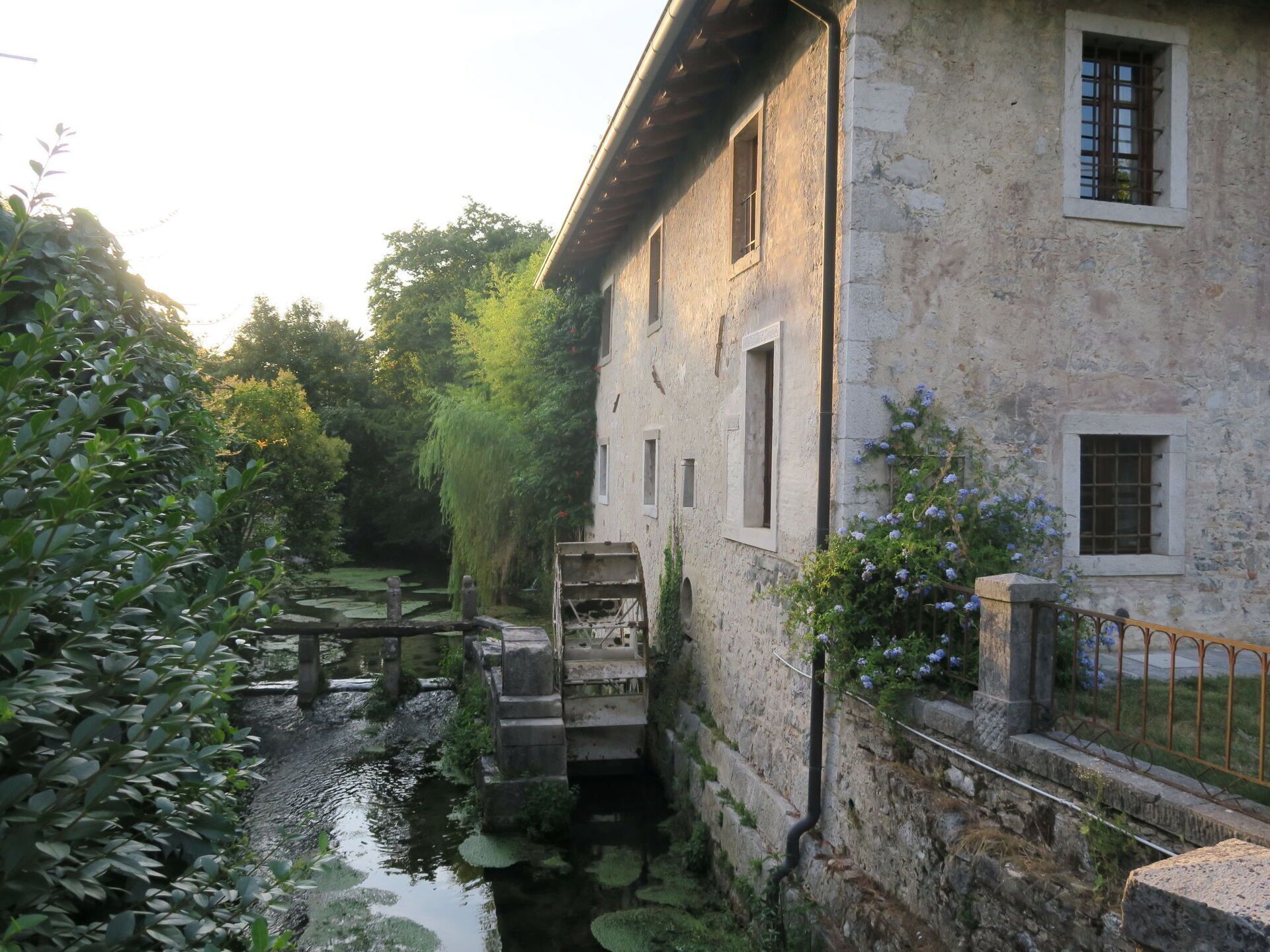 A stone building with multiple windows, a small bridge, and a pond with lily pads.