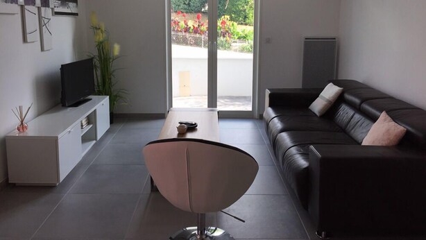Ein modernes Wohnzimmer mit einem dunkelbraunen Ledersofa, einem weißen Couchtisch, einem minimalistischen Sessel, einem Flachbildfernseher auf einem weißen Schrank und einer Glastür, die zu einem Garten führt.