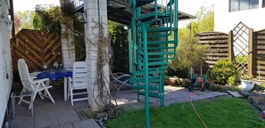 Eine Terrasse mit einem weißen Tisch und Stühlen, einer grünen Klettergerüst und einem Gartenschlauch.