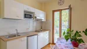 Una cocina compacta con armarios blancos, un microondas, un lavabo y un refrigerador pequeño. Hay una mesa de comedor con mantel floreado y una planta en maceta.