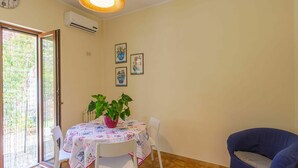 Un comedor con una mesa cubierta por un mantel floral, rodeada de sillas blancas. Hay una planta en maceta sobre la mesa, y se ve un sofá azul en la esquina.
