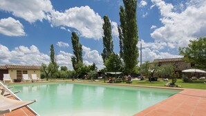 A rectangular swimming pool with a tiled deck, surrounded by a garden with trees and a building in the background.