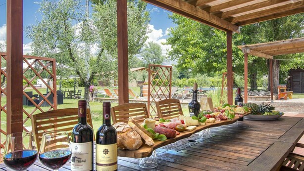 An outdoor dining area with wooden tables and chairs, a wooden pergola, and a view of a garden with trees and a lawn.