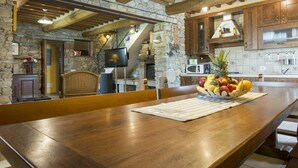 A rustic kitchen with stone walls, wooden beams, and a long wooden table with a fruit bowl.