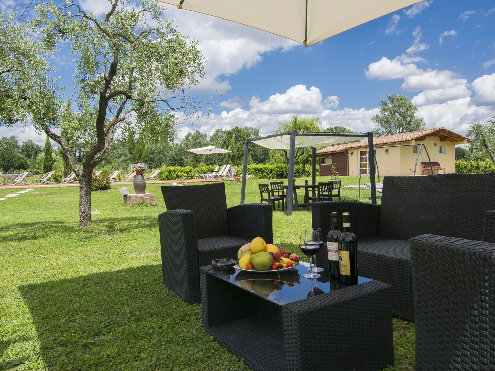 An outdoor seating area with wicker furniture, a table with fruit and wine bottles, and a pergola providing shade.