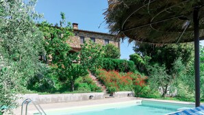 Ein Schwimmbecken mit Liegestühlen, im Hintergrund ein Steinhaus und üppiges Grün.