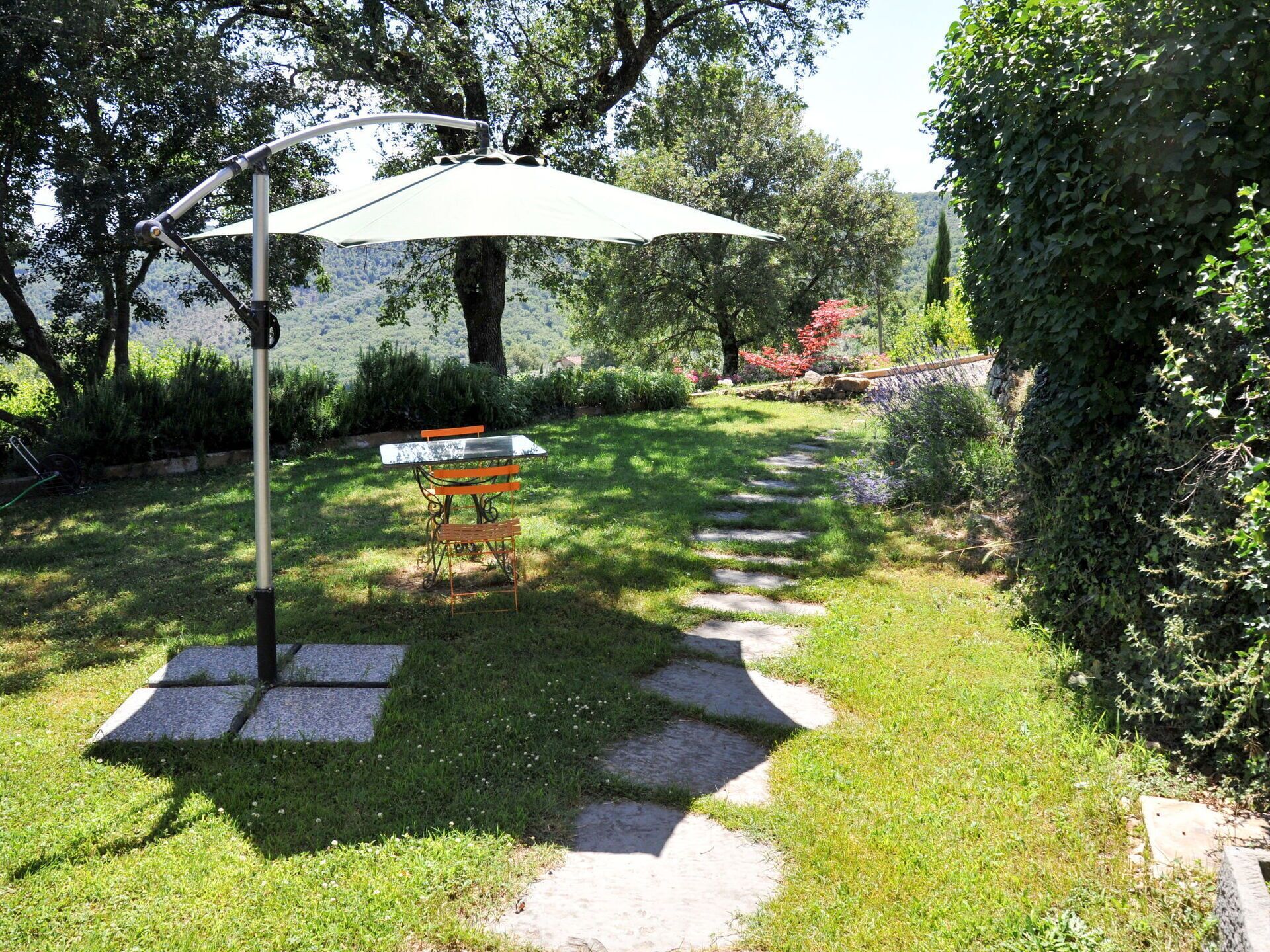 Un giardino con tavolo e sedie sotto un tetto riparatore, circondato da un vialetto in pietra e vegetazione lussureggiante.