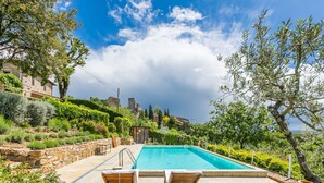 Zona de piscina con dos sillones de madera, un muro de piedra y una vista a un edificio histórico al fondo.