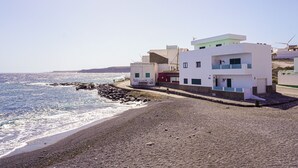 A coastal building with a beach view, a rocky shoreline, and a wind turbine in the background. - Las Eras - The House in front of the Sea (Arico)