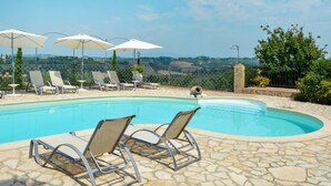 Zona de piscina con sillones de descanso y sombrillas, rodeada de baldosas de piedra y vistas al campo.