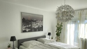 Een moderne slaapkamer met een groot bed, twee nachtkastjes met lampen, een stadslandschap schilderij aan de muur en dunne gordijnen.