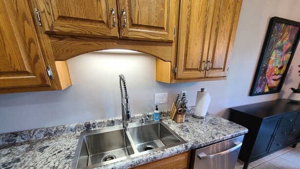 Una cocina con armarios de madera, pileta de acero inoxidable, lavavajillas y encimeras de granito.