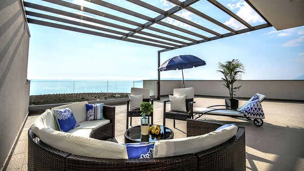 Een terras op het dak met uitzicht op de oceaan, ingericht met rieten meubels, een tafel met een vaas en een blauwe parasol.