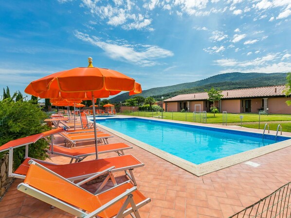 Une piscine entourée d’un patio en briques et d’un bâtiment doté d’un toit en tuiles, avec des chaises longues orange et des parasols.
