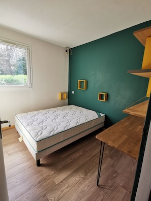 A bedroom with a bed, wooden floor, a window, and a green wall with three small shelves.